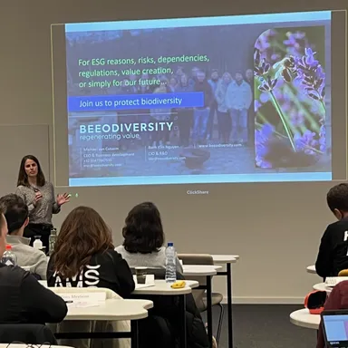 A speaker presents to participants seated at desks, in front of a projected slide reading “Join us to protect biodiversity”