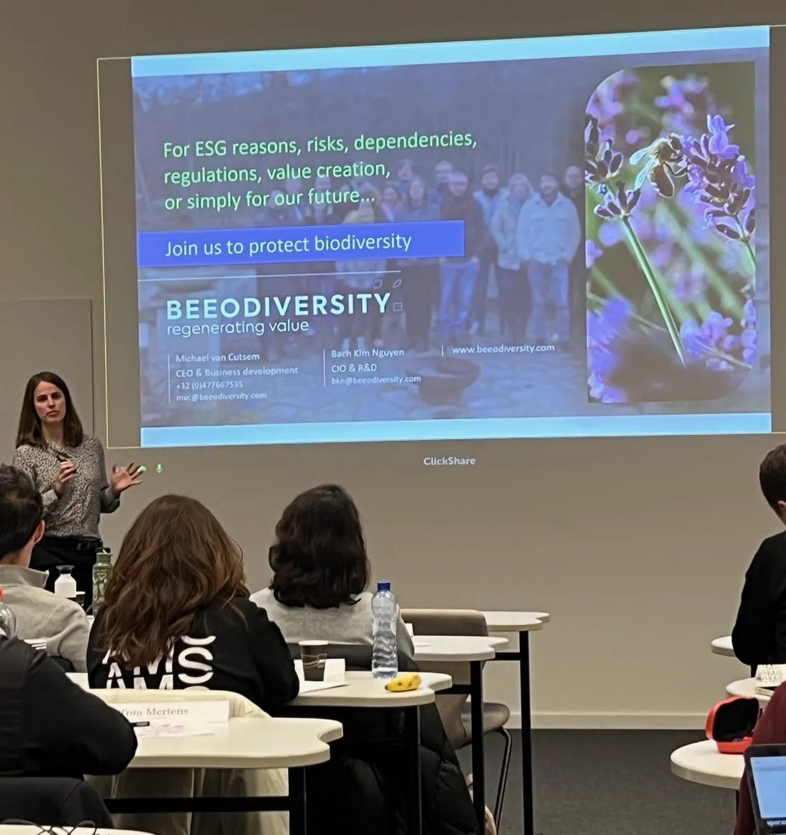 Un intervenant fait une présentation devant des participants assis à des tables, face à une diapositive projetée qui indique « Join us to protect biodiversity »