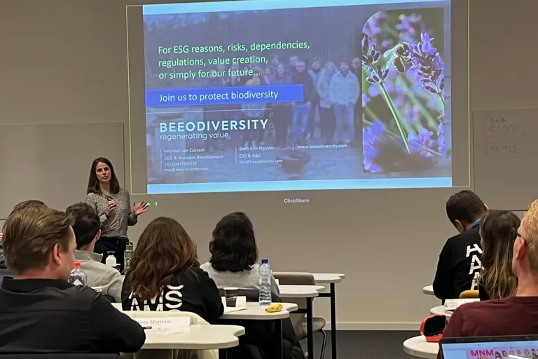 A speaker presents to participants seated at desks, in front of a projected slide reading “Join us to protect biodiversity”