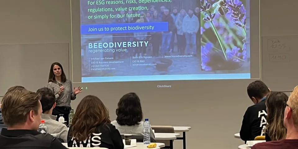 A speaker presents to participants seated at desks, in front of a projected slide reading “Join us to protect biodiversity”
